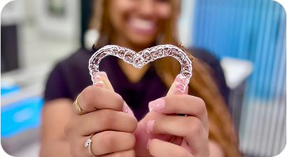 Stock image of a white woman with brown hair in her late 30s holding clear aligners for her teeth in her hand, smiling at the camera.